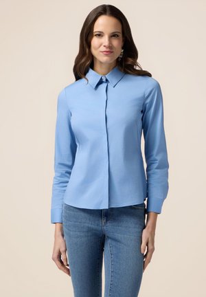 Camicia azzurro chiaro con maniche lunghe e bottone, collo classico, tessuto elegante e una vestibilità leggermente attillata, abbinata a jeans blu.
