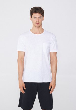 Weißes Baumwoll-T-Shirt mit rundem Ausschnitt und kurzen Ärmeln, ausgestattet mit einer linken Brusttasche, kombiniert mit schwarzen Shorts. Schlichtes Design und glatte Textur.