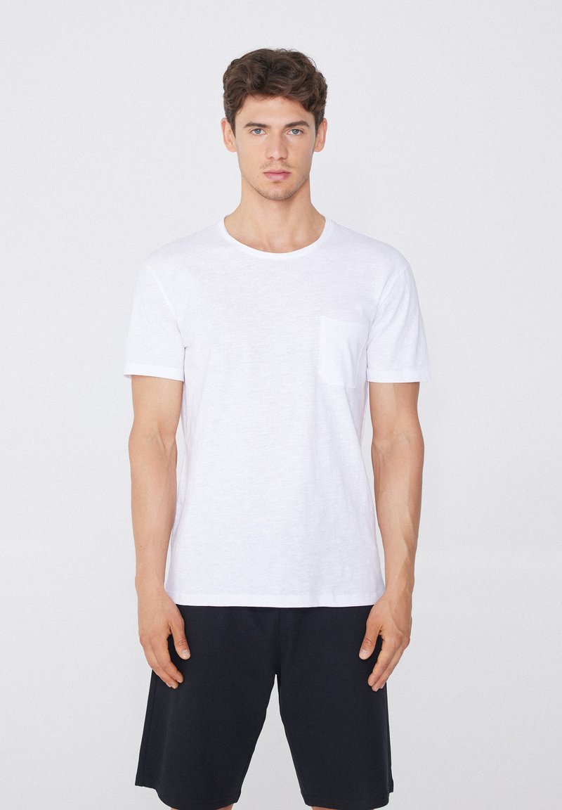 Camiseta blanca de algodón con cuello redondo y mangas cortas, que presenta un bolsillo en el pecho izquierdo, combinada con pantalones cortos negros. Diseño liso y textura suave.