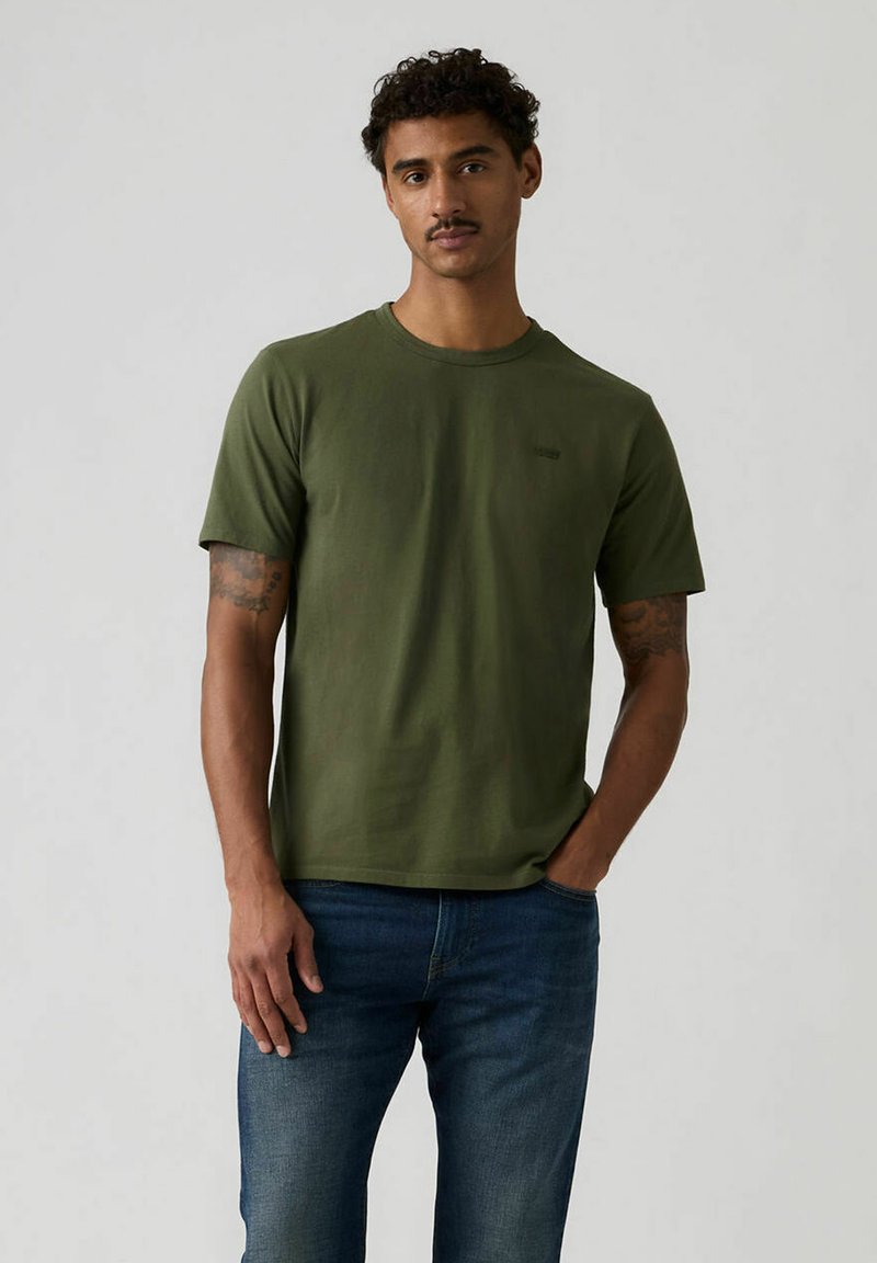 T-shirt en coton vert olive avec un col rond et des manches courtes. Présente un logo discret sur la poitrine et une texture lisse et uniforme. Porté avec un jean bleu.