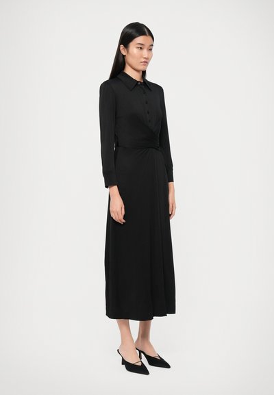 Rochie neagră tip wrap cu mâneci lungi, guler, nasturi în față și detaliu răsucit în talie; asortată cu pantofi negri cu vârf ascuțit și barete slingback.