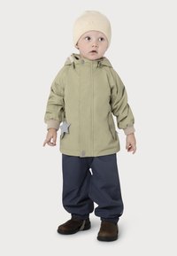 Kind in een lichtgroene, waterdichte jas met capuchon en steraccent, gecombineerd met donkerblauwe broeken en bruine laarzen. Crème gebreide beanie.