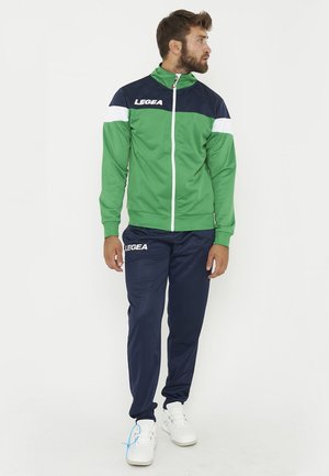 Homme debout portant un survêtement vert et bleu marine avec des chaussures blanches, regardant sur le côté sur un fond blanc uni.