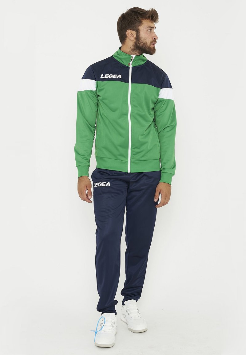 Homme debout portant un survêtement vert et bleu marine avec des chaussures blanches, regardant sur le côté sur un fond blanc uni.