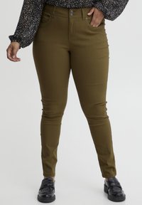 Olivno zeleni skinny jeans z gladko teksturo, ki imajo sprednje žepke, gumbasto zapiranje in ožji kroj, v kombinaciji s črnimi čevlji.