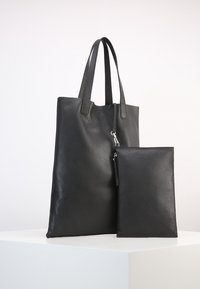 Sac fourre-tout en cuir noir avec surface texturée, double anse et pochette zippée amovible. Design simple, forme rectangulaire, accents minimalistes.