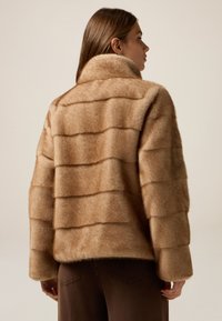 Chaqueta de piel sintética marrón con un alto cuello, que presenta un diseño acolchado a rayas. La textura es suave y esponjosa con un ajuste relajado.