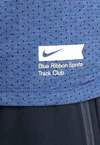 Niebieska tkanina o teksturze z małymi perforacjami, z białym logo Nike oraz napisem "Blue Ribbon Sports Track Club" nad czarnymi spodniami sportowymi.
