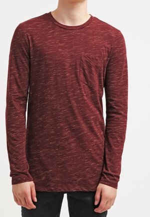 Person trägt ein dunkelrotes Langarmshirt mit einem dezenten weißen Sprenkelmuster und einer kleinen Brusttasche, kombiniert mit schwarzen Hosen.
