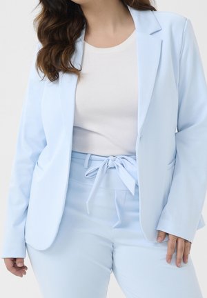 Žena v světle modrém blejzru a sladěných kalhotách s vysokým pasem a zavazovací páskem přes bílý žebrovaný top, stojící na jednobarevném pozadí.