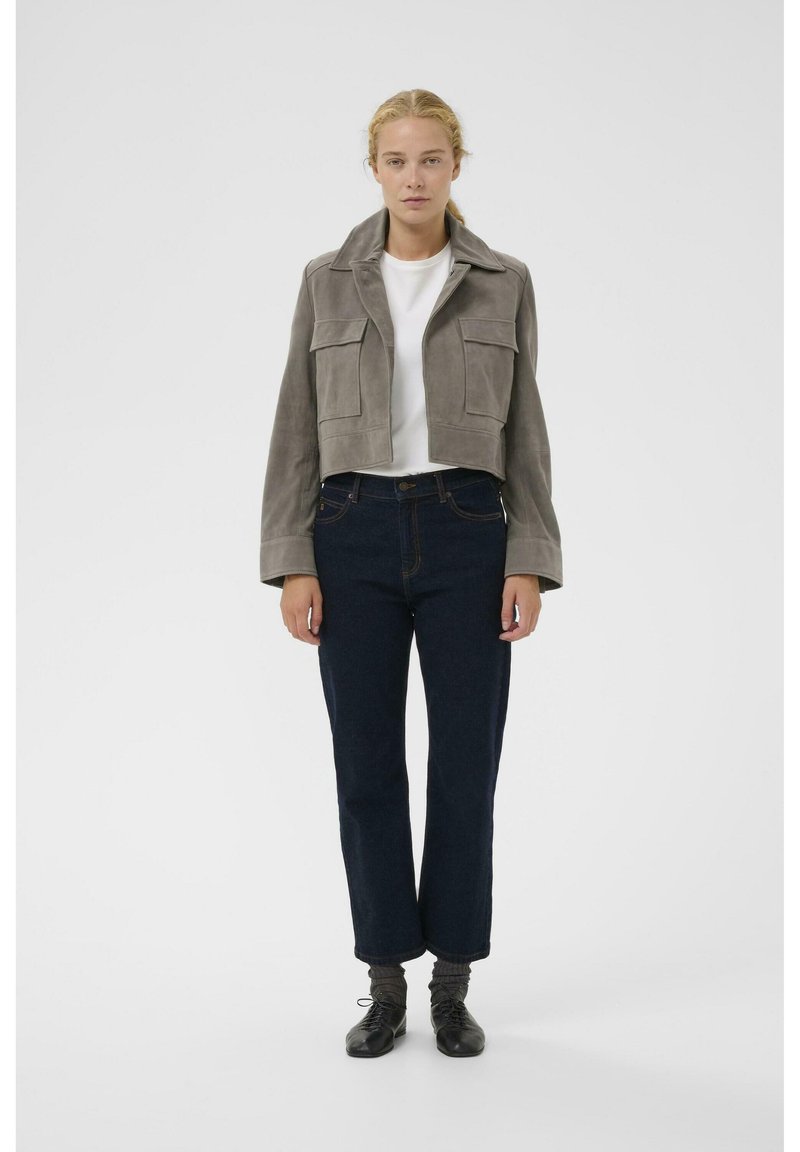 Pilkos spalvos zomšinė trumpa striukė su didelėmis kišenėmis su stačiakampių atvartais, derinama su tamsiai mėlynomis tiesių kojų džinsais ir juodais batais su pilkomis ribinėmis kojinėmis.