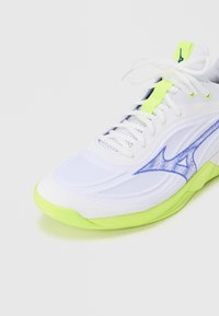 Άσπρο αθλητικό παπούτσι με σόλα και λεπτομέρειες σε νέον πράσινο, μπλε λογότυπο στο πλάι και άσπρα κορδόνια δεμένα σε φιόγκο σε απλό φόντο.