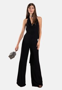 Schwarzes Halterneck-Top mit drapiertem Design, kombiniert mit weit geschnittenen Hosen. Begleitet von einem silberfarbenen Choker und einer kleinen grauen Clutch.