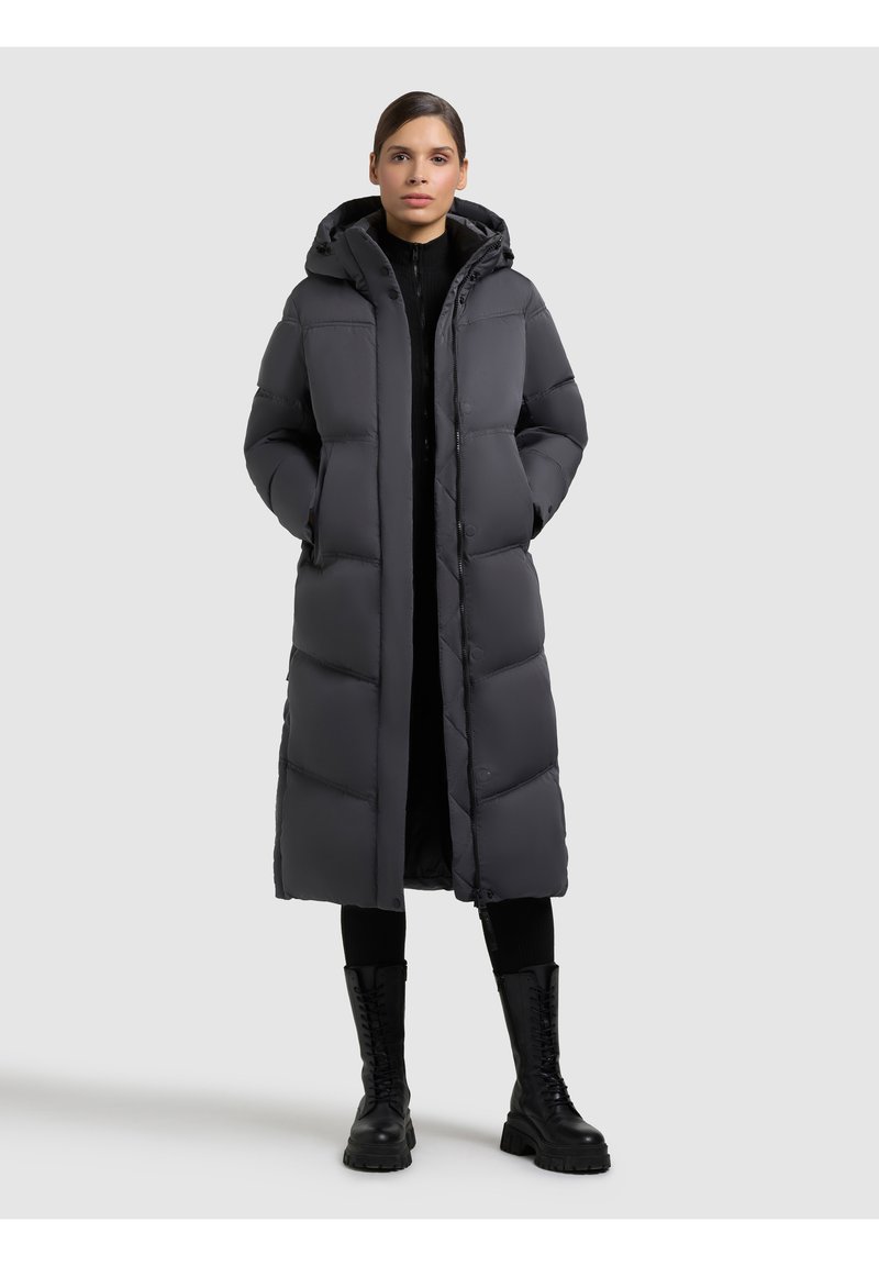 Geacă puf gri cu glugă, având un design lung și drept, textură matlasată, nasturi tip snap și buzunare laterale. Purtată peste o ținută neagră.