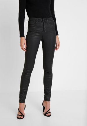 Femme portant un pantalon en cuir noir skinny taille haute, un haut noir à manches longues, et des sandales à talons hauts noires avec des lanières, debout sur un sol blanc.