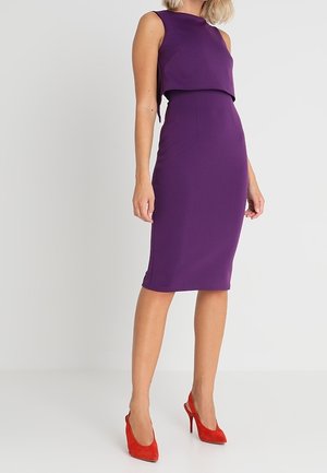 Žena v fialových midi šatech bez rukávů a červených lodičkách, stojící před jednobarevným světle šedým pozadím.