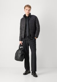 Giacca imbottita nera con trapuntatura orizzontale, abbinata a una camicia nera e cravatta. Indossa pantaloni e scarpe neri, mentre tiene una borsa nera.