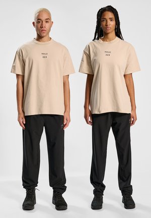 Beige katoenen T-shirts met "HALO 223" in het zwart gedrukt, gecombineerd met zwarte broeken en zwarte laarzen. Modellen tonen een relaxte pasvorm en een minimalistisch ontwerp.