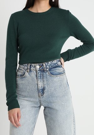 Sweater vert en tricot à manches longues avec un col rond, associé à un jean en denim bleu clair taille haute avec de légers effets usés.