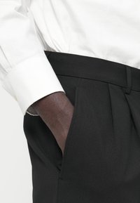 Μαύρο παντελόνι κατά παραγγελία με πιέτες και πλαϊνή τσέπη, συνδυασμένο με λευκό πουκάμισο μακρυμάνικο. Το ύφασμα έχει λεία υφή.