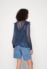 Blusa de gasa azul marino con detalles rizados, patrón de lunares y abertura en la parte posterior, combinada con shorts de mezclilla azul claro.