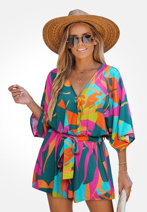 Mujer con un vibrante mono multicolor de mangas anchas, un sombrero de paja, gafas de sol, collares dorados y sosteniendo un bolso de mano tejido.