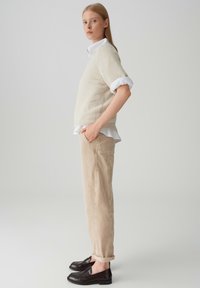 Maglione lavorato a maglia beige con maniche corte sopra una camicia bianca con bottoni, abbinato a pantaloni beige e mocassini scuri. Fit semplice e rilassato.
