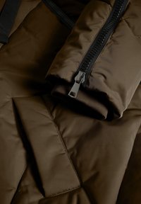 Brązowa kurtka puchowa z detalami, takimi jak czarny zamek błyskawiczny, teksturowana tkanina oraz złożony mankiet z jednolitym wnętrzem.