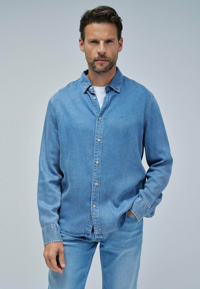 Camisa de mezclilla azul claro con diseño de botones, mangas largas y cuello. Presenta un logo en el pecho y se muestra con pantalones de mezclilla a juego.