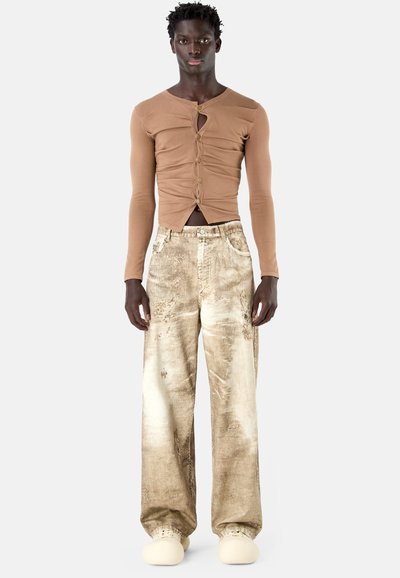 Homme portant un haut beige à manches longues, froncé, boutonné, avec une découpe à l'avant, un pantalon beige texturé à jambes larges et des chaussures couleur crème.