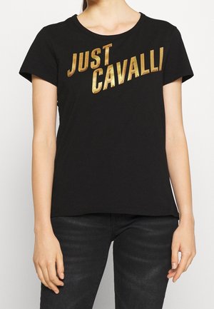 Kobieta w czarnej koszulce z krótkim rękawem, z dużym złotym napisem "JUST CAVALLI" na piersi.