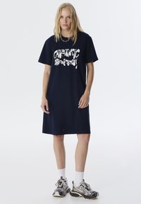 Navyblauwe katoenen t-shirt jurk met vetgedrukte witte grafische tekst, korte mouwen en een rechte snit. Gecombineerd met chunky sneakers en witte sokken.