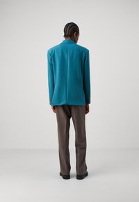 Türkiser Blazer mit taillierter Passform, einem Rückenschlitz und Knopfdetails. Kombiniert mit hellbraunen Hosen und schwarzen Schuhen. Glatte Stofftextur.