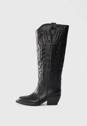 Des bottes de cowboy en cuir noir présentent une tige haute avec des motifs de couture en relief et un bout pointu. Le talon est légèrement incliné et épais.