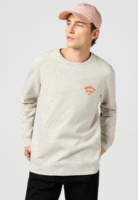 Sudadera gris claro con mangas largas, cuello redondo y logo naranja "Wrangler Jeans 1947" en el pecho. Lleva puestas unos pantalones negros y una gorra rosa.