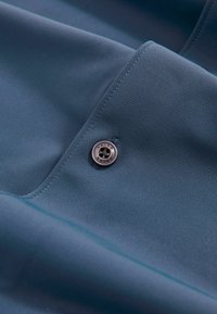 Texturiertes blaues Gewebe mit glatter Oberfläche und einem silbernen Knopf mit einem Design aus vier Löchern, der an einer Naht befestigt ist.