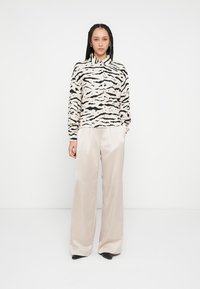Chemise à manches longues à boutons beige avec motif rayures tigre noires, associée à un pantalon large beige clair lisse et des talons noirs.
