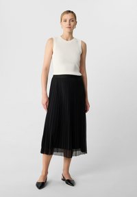 Zwarte plissé midi-rok met een doorzichtige zoom, gecombineerd met een witte geribbelde mouwloze top. De schoenen zijn zwarte slingbacks met een spitse neus.