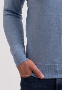 Vaaleansininen neulepusero, jossa on pitkät hihat ja ribattu pääntie, jonka pinta on sileä ja hieman vartalonmyötäinen muoto.