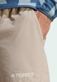 Bež kratke hlače Terrex s elegantnim dizajnom, koje imaju bočnu džepu i bijeli logo. Tkanina ima glatku teksturu i lagani izgled.
