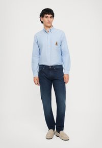 Cămașă bleu deschis cu manșete, dotată cu un logo mic pe piept, asortată cu blugi bleumarin și mocasini bej. Vedere din întreaga corp pe un fundal neutru.