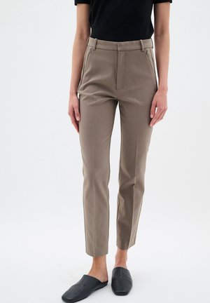 Kobieta ubrana w dopasowane spodnie w kolorze taupe, czarny krótki top i czarne buty wsuwane, stojąca na jednolitym tle.