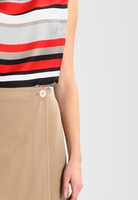 Mouwloos topje met brede rode, zwarte, beige en witte strepen; gecombineerd met een knielange tan kleurige rok met een knoopdetail.