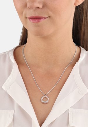 Silver necklace with a circular pendant featuring a rose gold accent, adorned with small clear crystals, worn over a white blouse.