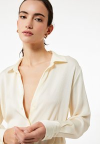 Krämfärgad blus i ett lätt, texturerat tyg med djup V-ringning, lång ärm och knapprygg för stängning.
