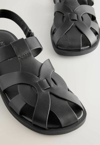 Sandales pour homme en cuir noir avec design à lanières tissées et sangle de cheville réglable sur une surface claire.