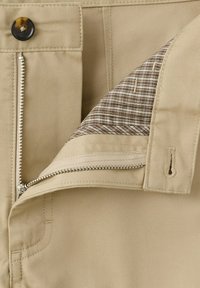 Gros plan sur la taille d'un pantalon beige avec une fermeture éclair métallique partiellement ouverte, bouton fermé et doublure intérieure en tissu à carreaux visible.
