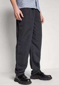 Schwarze Hosen mit lockerem Schnitt, aus glattem Material. Kombiniert mit schwarzen klobigen Schuhen. Das Outfit hat einen lässigen Look mit minimalen Details.