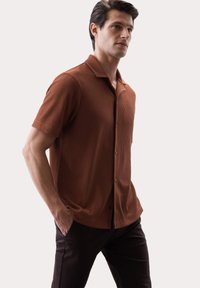 Chemise à manches courtes, côtelée, de couleur marron foncé avec col et fermeture à boutons, assortie à un pantalon foncé, sur un fond neutre.