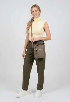 Frau trägt einen ärmellosen gelben Rollkragenpullover, olivgrüne Hose, weiße Turnschuhe und eine braune Umhängetasche mit mehreren Reißverschlusstaschen.